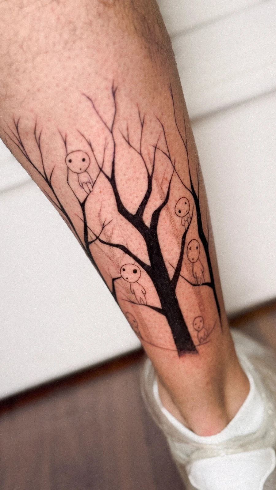 Tatuaje de un árbol sin hojas con pequeños espíritus del bosque (kodamas, de La princesa Mononoke) en la pantorrilla